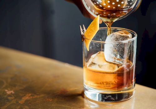 A drink being poured into a glass with ice and a lemon twist garnish on a table.