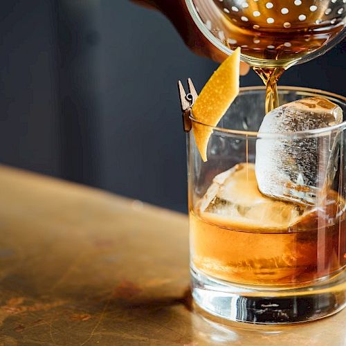 A drink being poured into a glass with ice and a lemon twist garnish on a table.