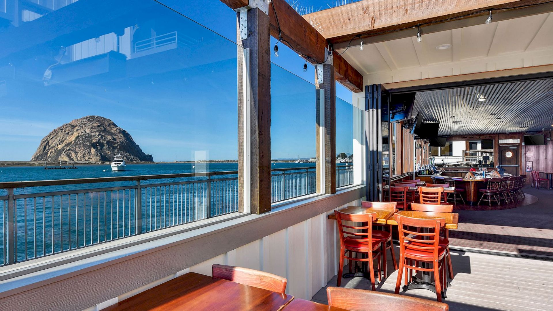 A restaurant with wooden tables, ocean view, and a large rock formation in the distance through glass windows.