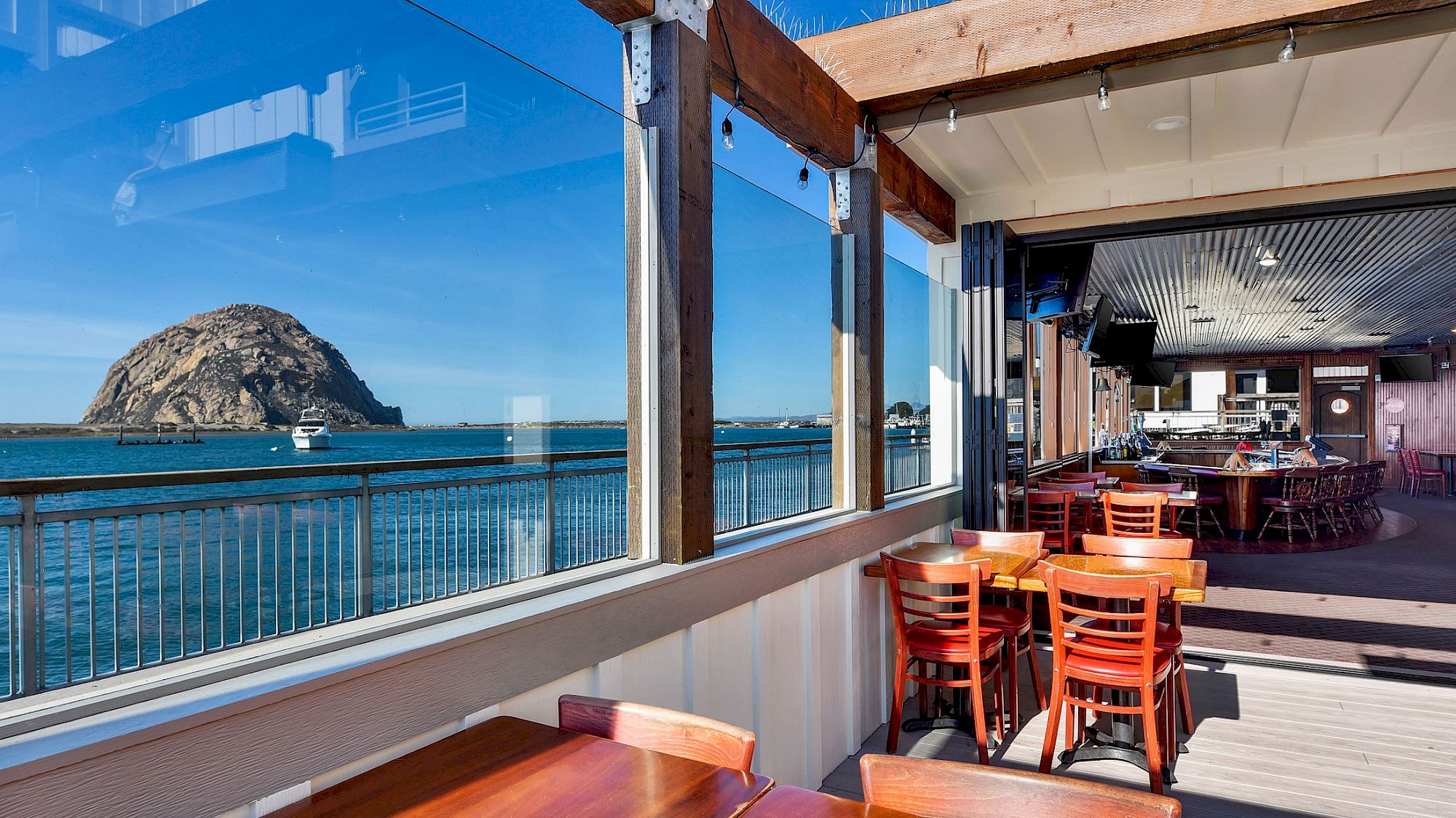 A restaurant with wooden tables, ocean view, and a large rock formation in the distance through glass windows.