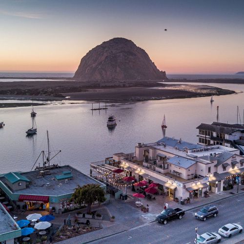 A coastal town at sunset with boats in the water and a large rock formation in the background, featuring waterfront buildings and street activity.