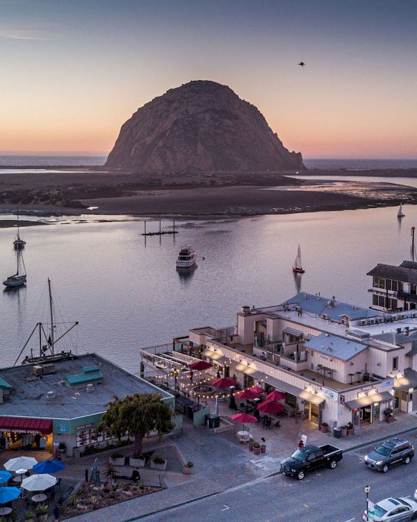 A coastal town at sunset with boats in the water and a large rock formation in the background, featuring waterfront buildings and street activity.