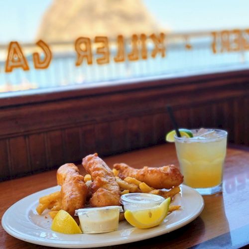 The image shows a plate of fish and chips with lemon wedges and dipping sauces, accompanied by a drink with a lime slice, on a table by a window.