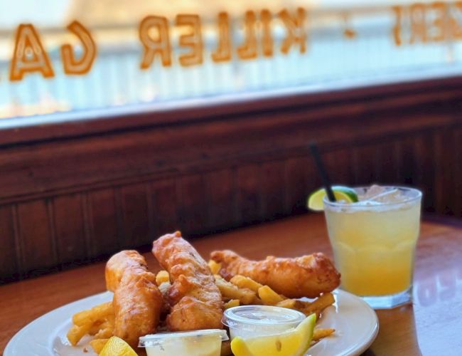 The image shows a plate of fish and chips with lemon wedges and dipping sauces, accompanied by a drink with a lime slice, on a table by a window.