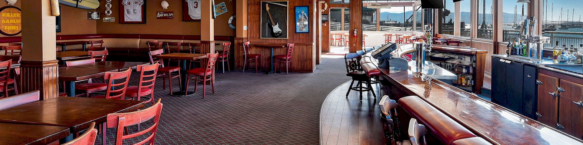 A sports bar with red chairs, tables, bar counter, sports memorabilia on walls, and a view of a marina through large windows.