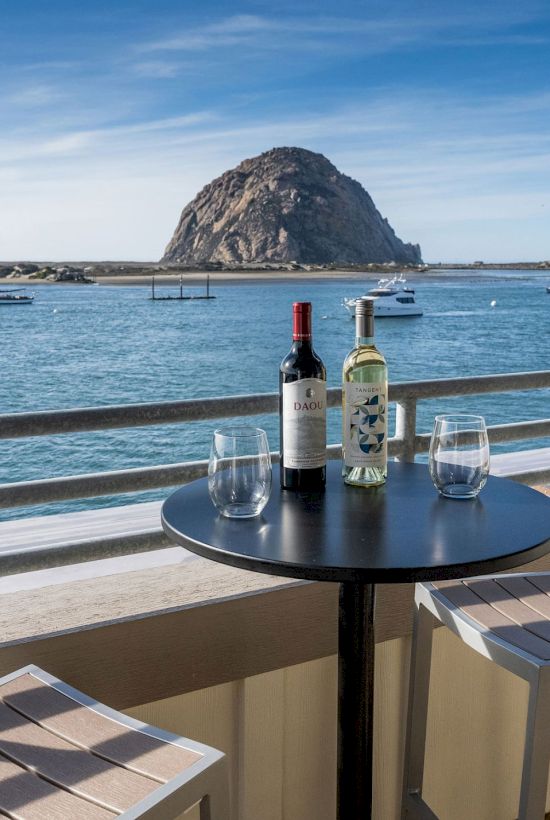 A scenic view shows two bottles of wine and glasses on a table with chairs, overlooking the ocean and a large rock formation in the background.