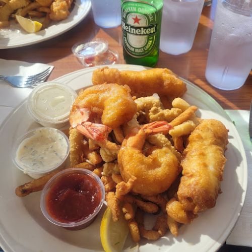 A plate of fried seafood with shrimp, fish, and French fries, accompanied by dipping sauces. A lemon wedge and a Heineken are nearby.