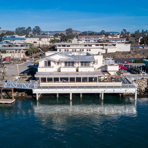A large waterfront building on stilts over the water, surrounded by other buildings and a clear blue sky.