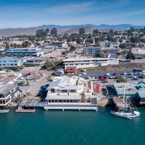 A coastal town features waterfront buildings, calm turquoise waters, and a small docked sailboat under a clear blue sky.
