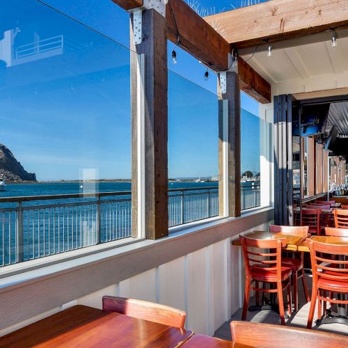 A dining area with wooden tables and chairs overlooks a large rock formation by the ocean through glass windows.