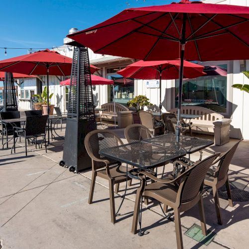 Outdoor patio with red umbrellas, tables, chairs, and a heater. Potted plants and lights add to the setting near a waterfront location.