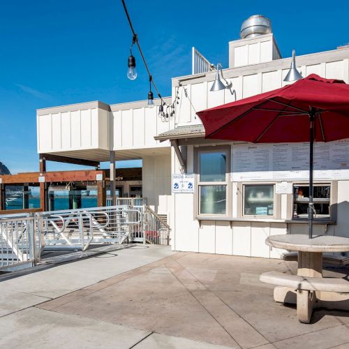 A waterfront eatery with a red umbrella, picnic table, and view of a large rock formation under a clear blue sky.