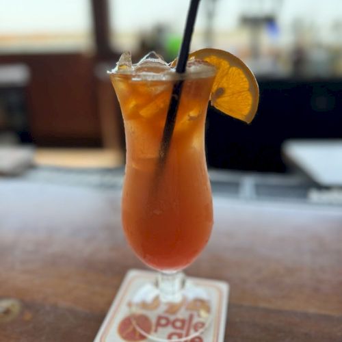A cocktail garnished with an orange slice and a straw sits on a coaster at a bar.