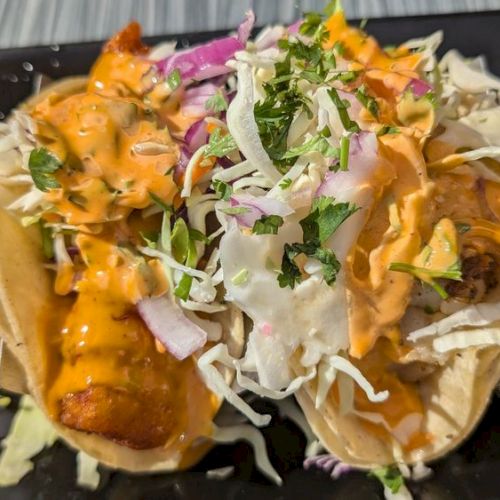 Two tacos with crispy filling, topped with shredded cabbage, sauce, diced onions, and cilantro, served on a black rectangular plate.