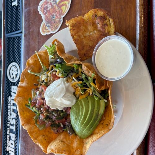 A taco salad in a tortilla bowl with avocado, sour cream, and dressing, served on a wooden table with a drink coaster nearby.
