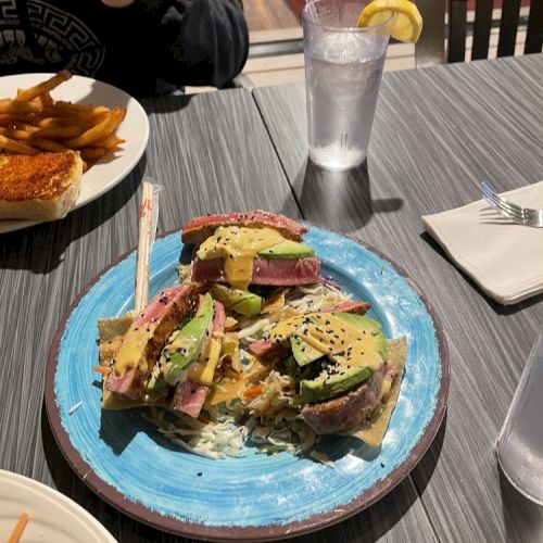 A colorful dish with layered food on a blue plate, a glass of water with lemon, and another plate with food beside it.