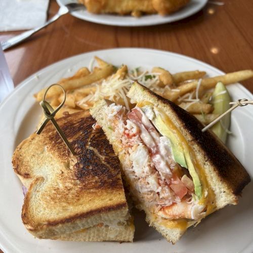 A sandwich with fries on a plate, filled with lettuce, tomato, and possibly seafood. Another plate with fried food is in the background.