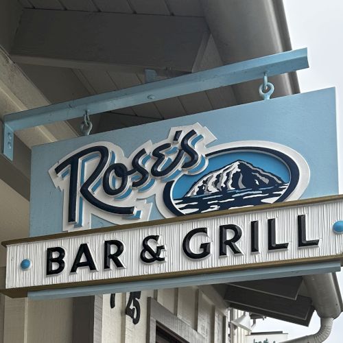 The image shows a sign for "Rose's Bar & Grill" featuring a mountain graphic, hanging outside a building.