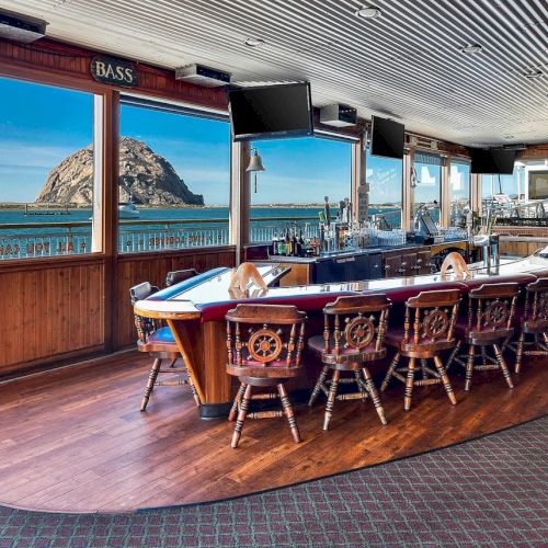 The image shows a boat-shaped bar with wooden chairs inside a room overlooking the ocean and a large rock formation through the windows.