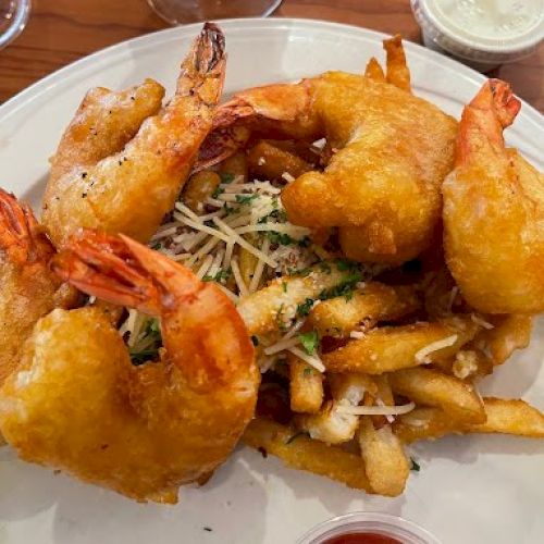 A plate with battered fried shrimp on top of fries, sprinkled with cheese and herbs, with dipping sauces on the side.