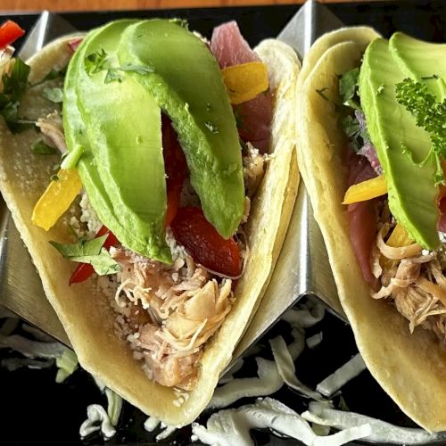 Three tacos filled with shredded meat, avocado slices, colorful bell peppers, and herbs on a black tray with lemon wedges.
