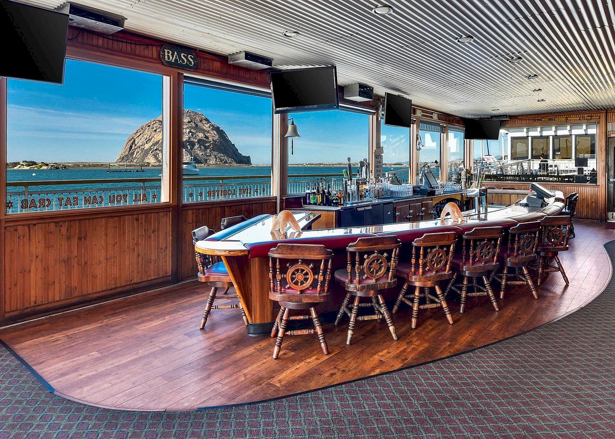 The image shows a boat-themed bar with a view of a large rock formation through the windows, featuring wooden nautical decor and seating.