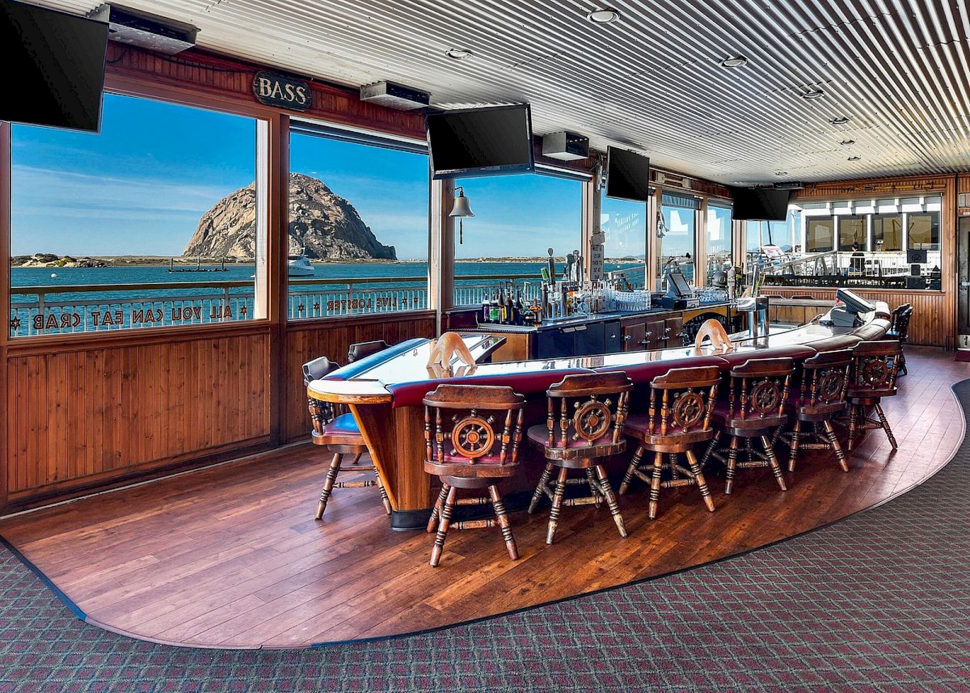 The image shows a boat-themed bar with a view of a large rock formation through the windows, featuring wooden nautical decor and seating.