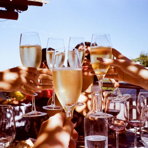 People are raising champagne glasses in a toast, with sunlight casting shadows on a table filled with glassware outdoors.