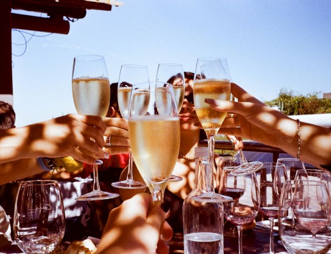 People are raising champagne glasses in a toast, with sunlight casting shadows on a table filled with glassware outdoors.