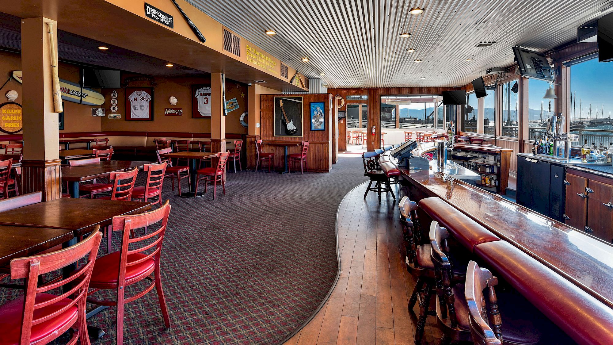 The image shows an empty sports bar with red chairs, wooden tables, a bar counter, sports memorabilia on the walls, and windows with a marina view.